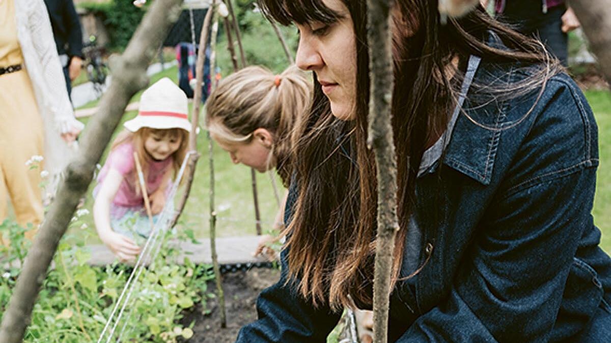 Außenaufnahme: Junge Frau hockt neben einem Beet, im Hintergrund ein Kind