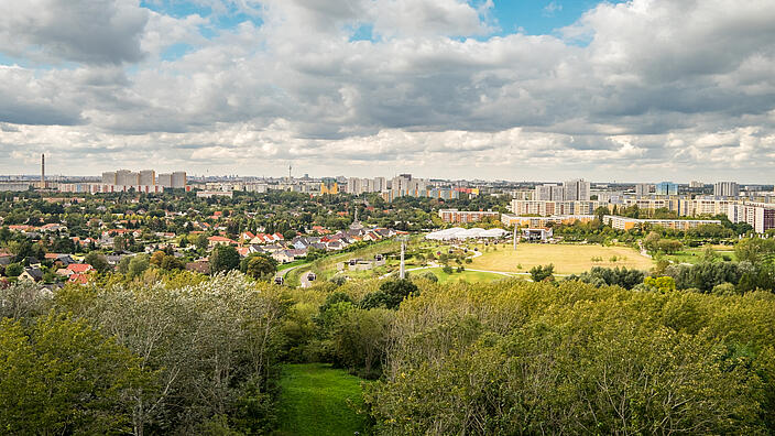 Die Außenaufnahme zeigt eine Panoramaaussicht über den Berliner Bezirk Marzahn-Hellersdorf vom Kienberg aus. Über den zahlreichen Wohnhäusern sammeln sich Wolken am Himmel. Zwischen den Häuserreihen befinden sich viele grüne Bäume. Ganz in der Ferne kann man das berühmte Berliner Wahrzeichen, den Fernsehturm, erspähen.