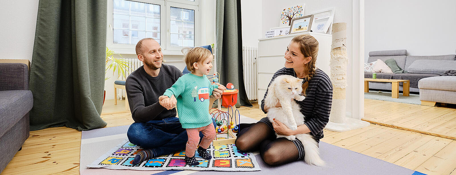 Innenaufnahme: Junger Mann und junge Frau sitzen mit Kleinkind und weißer Katze auf einem Teppich im Wohnzimmer