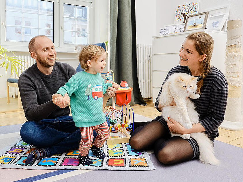 Innenaufnahme: Junger Mann und junge Frau sitzen mit Kleinkind und weißer Katze auf einem Teppich im Wohnzimmer