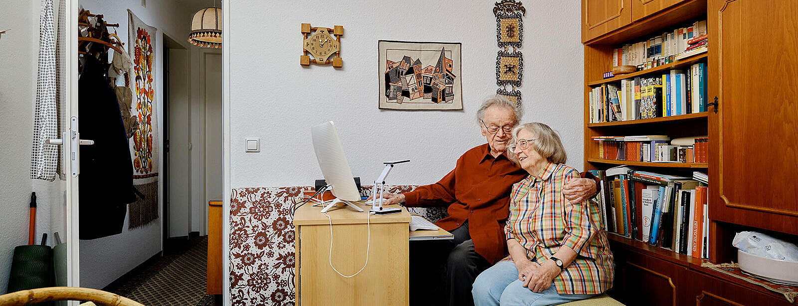 Innenaufnahme: Älteres Paar sitzt vor einem Schreibtisch und umarmt sich, die Frau schaut auf einen Computermonitor