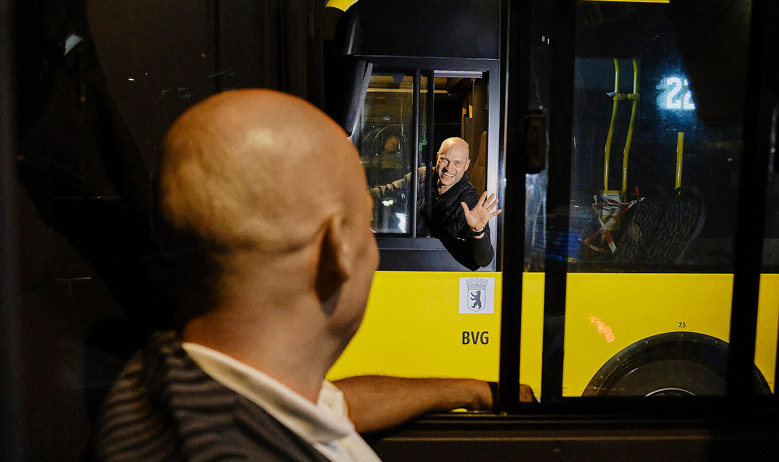 Außenaufnahme: Mann am Steuer eines gelben BVG-Busses grüßt anderen Mann