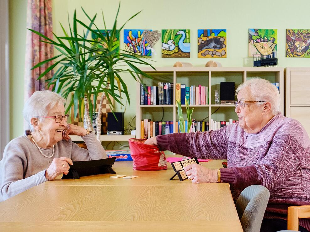 Innenaufnahme: Zwei ältere Damen sitzen gegenüber an einem Tisch und spielen ein Spiel, seitlich steht ein Rollator
