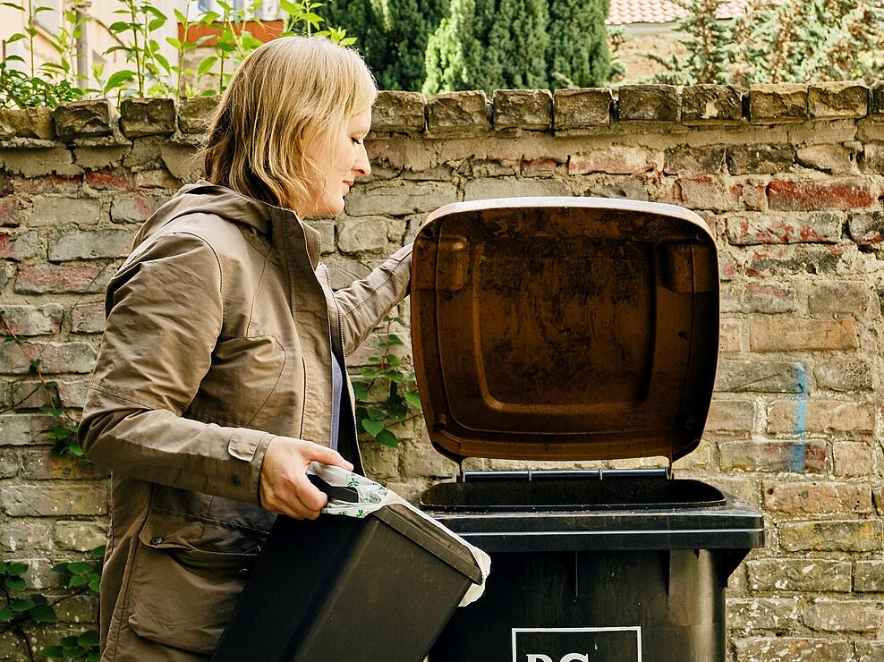 Außenaufnahme: Frau mit Mülleimer in der Hand steht vor aufgeklappter Biogut-Tonne