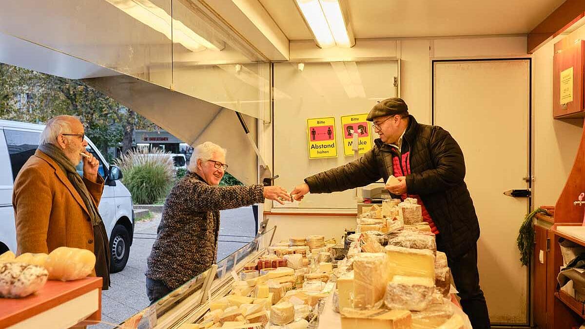 Außenaufnahme: Zwei ältere Männer an einem Käsestand, Verkäufer reicht etwas über die Theke