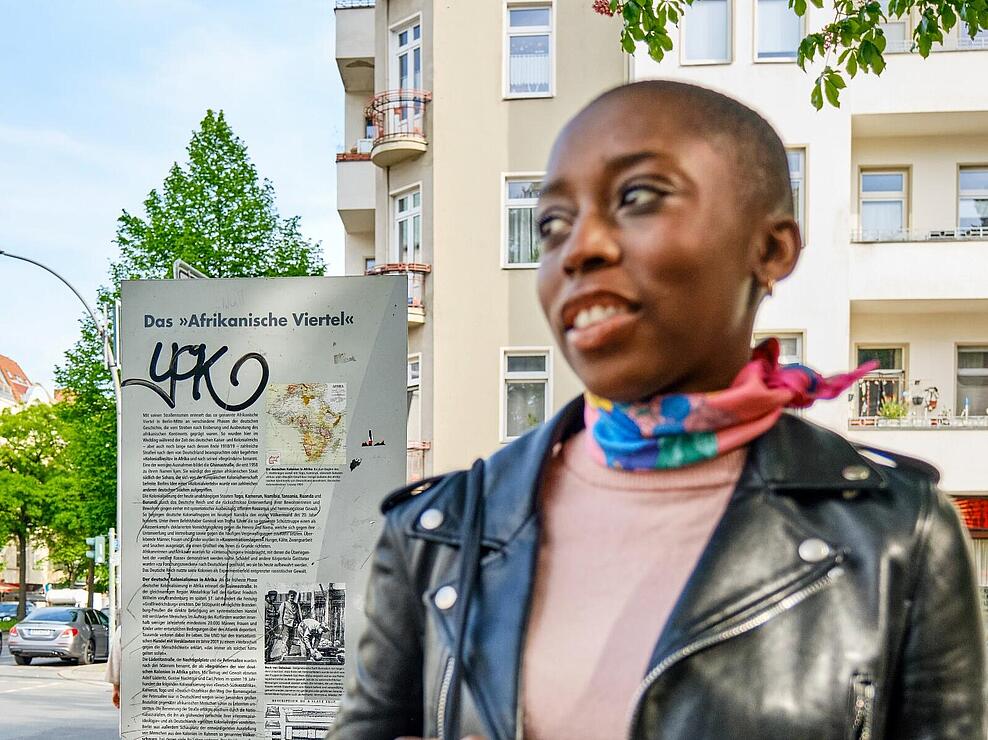 Außenaufnahme: Afrikanisch gelesene Frau auf dem Bürgersteig, im Hintergrund Infostele zum Afrikanischen Viertel
