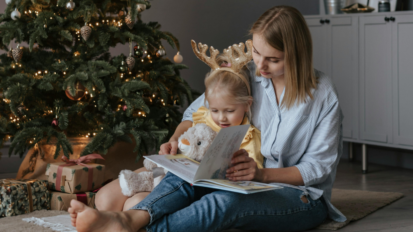 Mutter und Tochter sitzen neben einem geschmückten Weihnachtsbaum auf dem Boden und blättern in einem Kinderbuch.