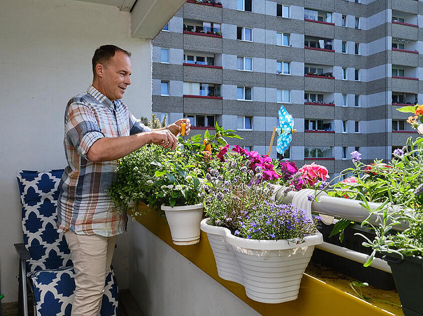 Außenaufnahme: Mann steht auf einem Balkon mit Grünpflanzen und Blumen, im Hintergrund eine Hochhausfassade