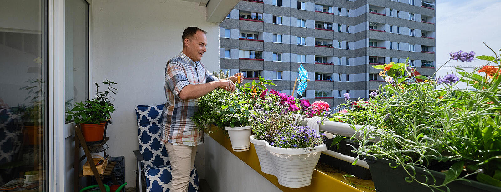 Außenaufnahme: Mann steht auf einem Balkon mit Grünpflanzen und Blumen, im Hintergrund eine Hochhausfassade