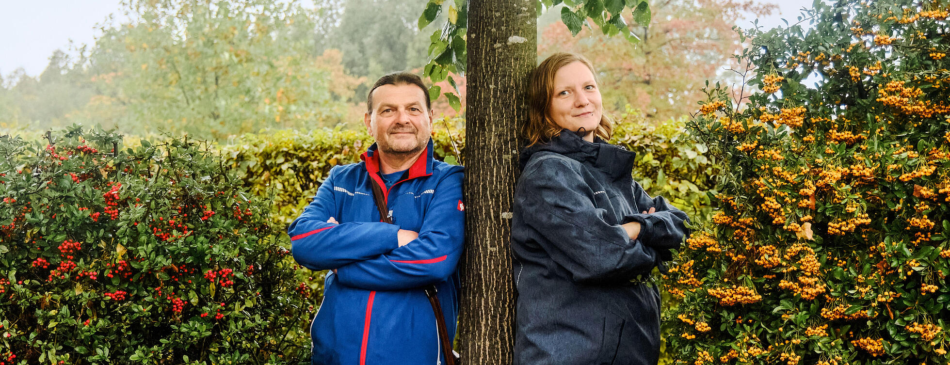 Außenaufnahme: Mann und Frau lehnen sich an Baum, daneben Hecken