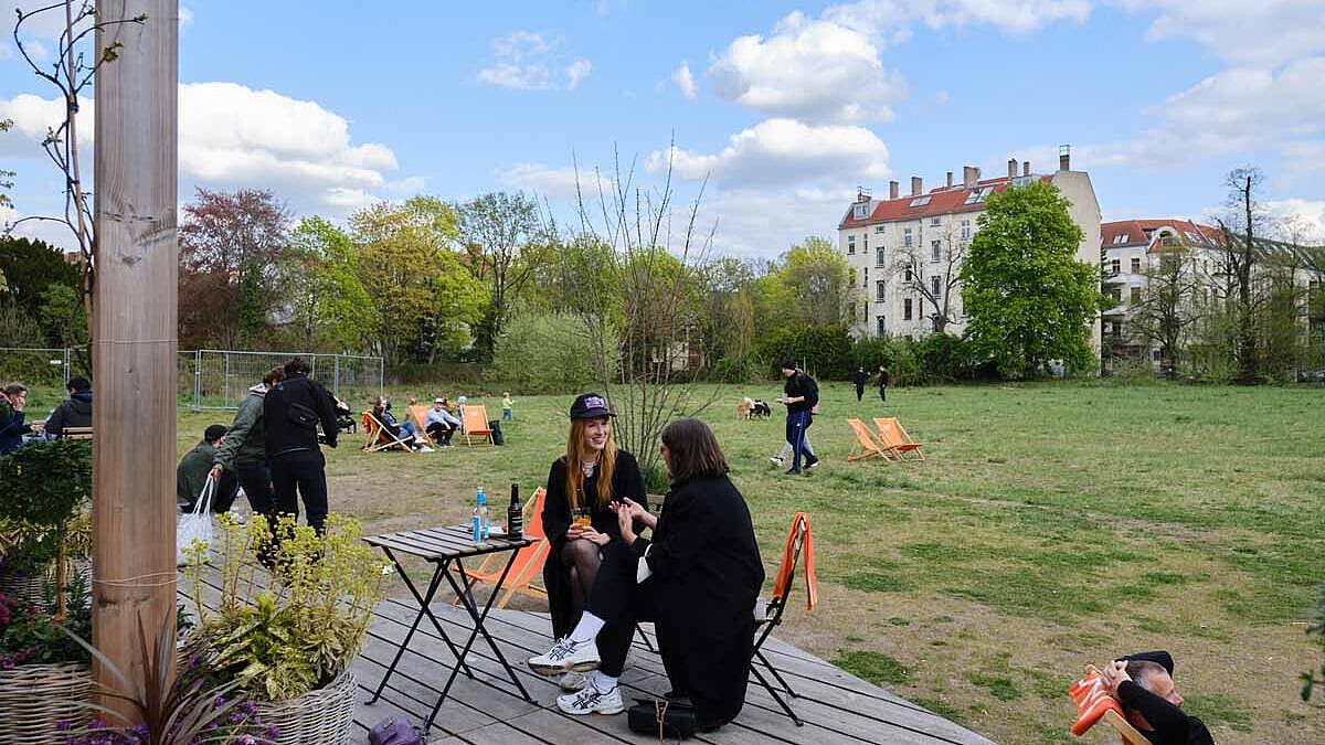 Außenaufnahme: Zwei Frauen sitzend auf Holzterrasse, neben ihnen auf einem Tisch Getränke, im Hintergrund Grünfläche