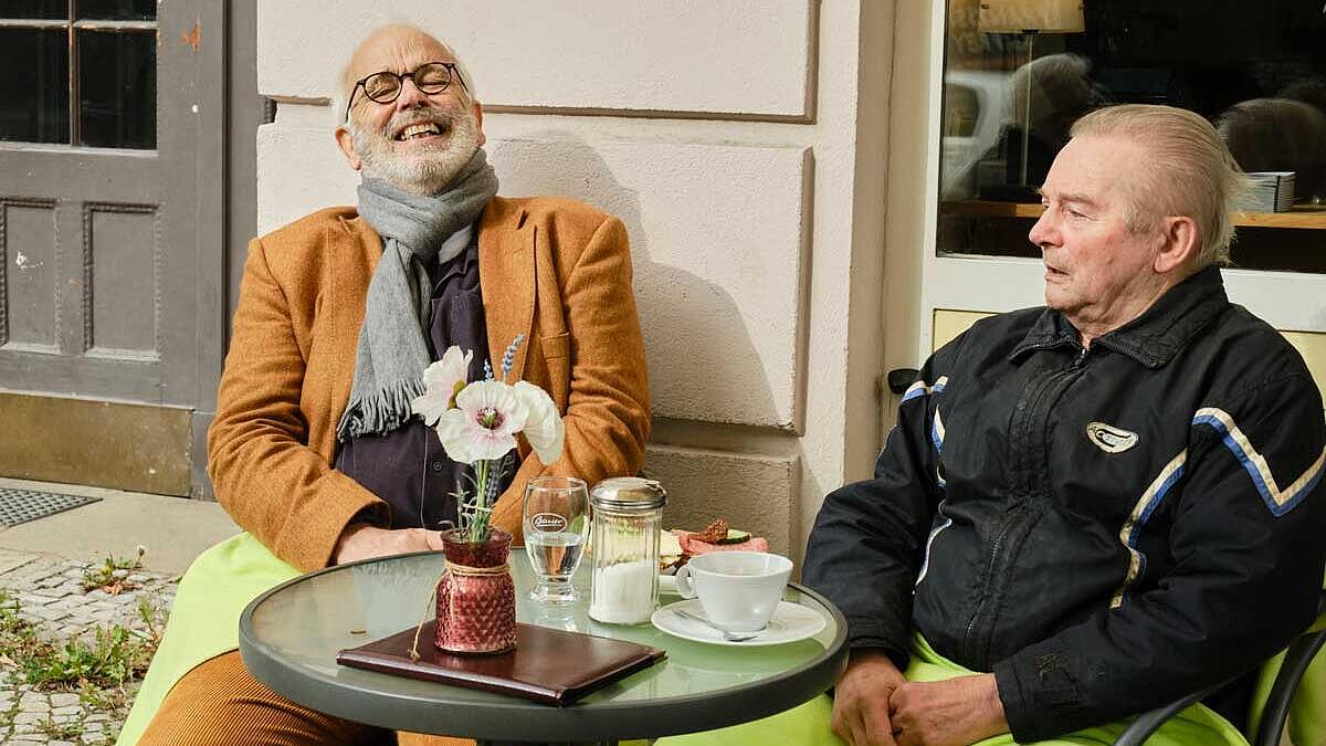 Außenaufnahme: Zwei ältere Männer sitzen an einem runden Café-Tisch, vor sich Getränke