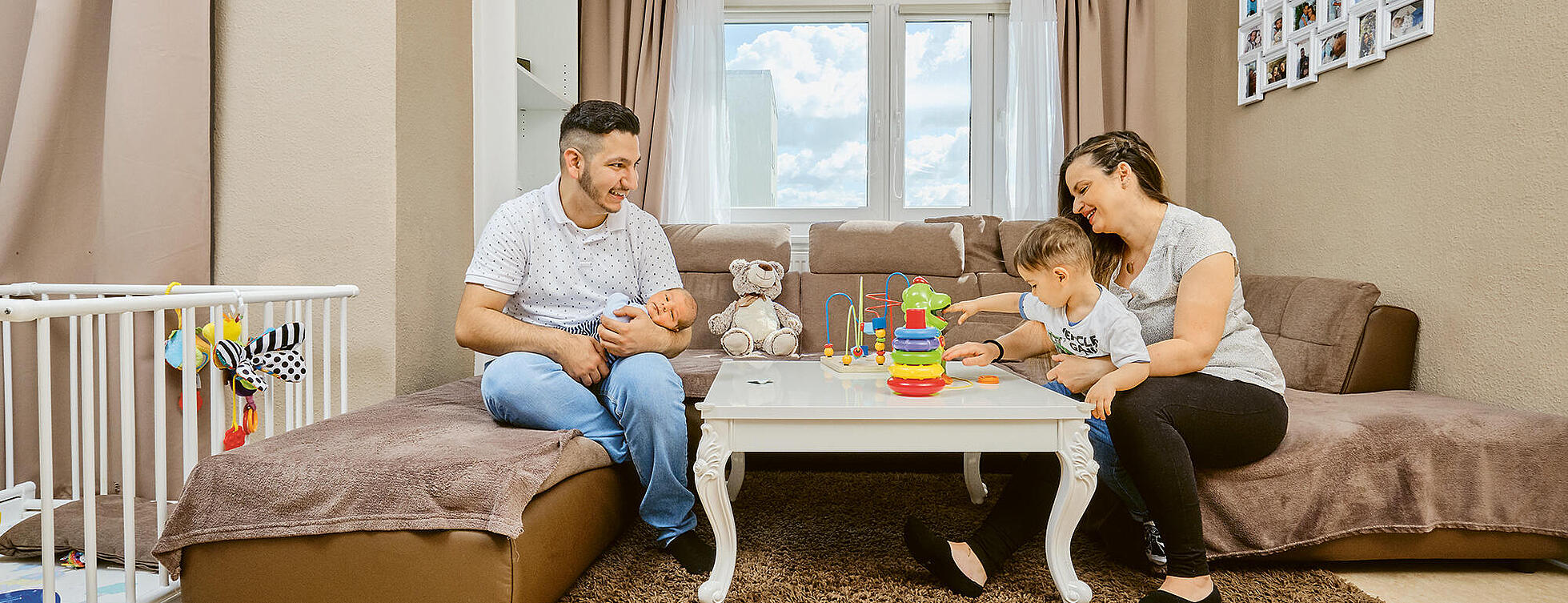 Innenaufnahme: Mann und Frau sitzen mit Baby und Kleinkind auf einer Couch, seitlich steht ein Babybett