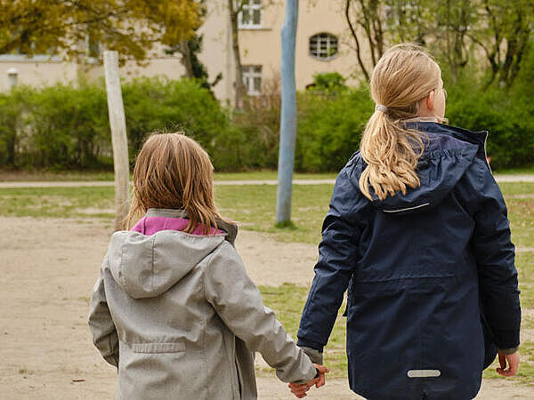 Außenaufnahme: Zwei Mädchen, von hinten aufgenommen, halten sich an den Händen