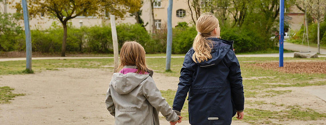 Außenaufnahme: Zwei Mädchen, von hinten aufgenommen, halten sich an den Händen