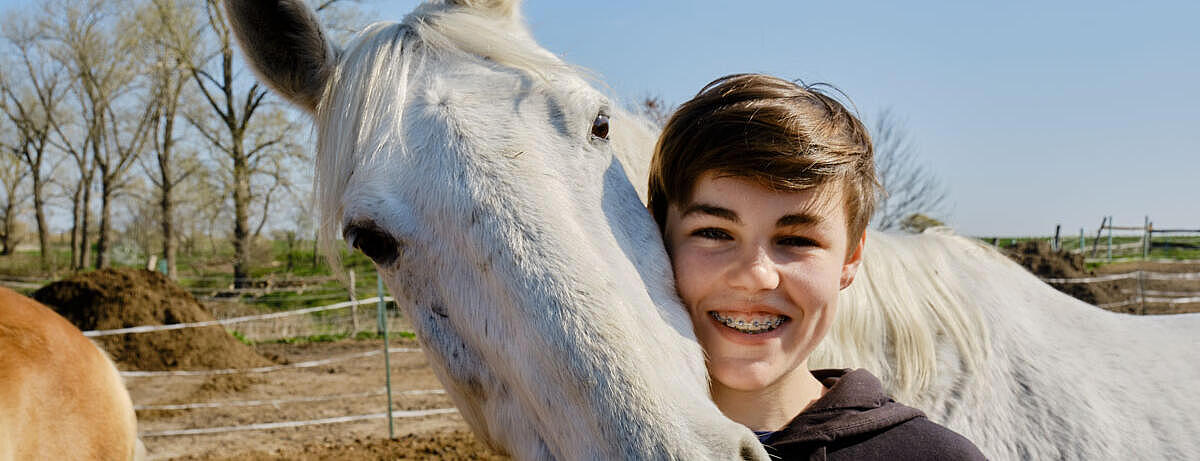 Außenaufnahme: Weißes Pferd legt Kopf auf die Schulter eines Jungen