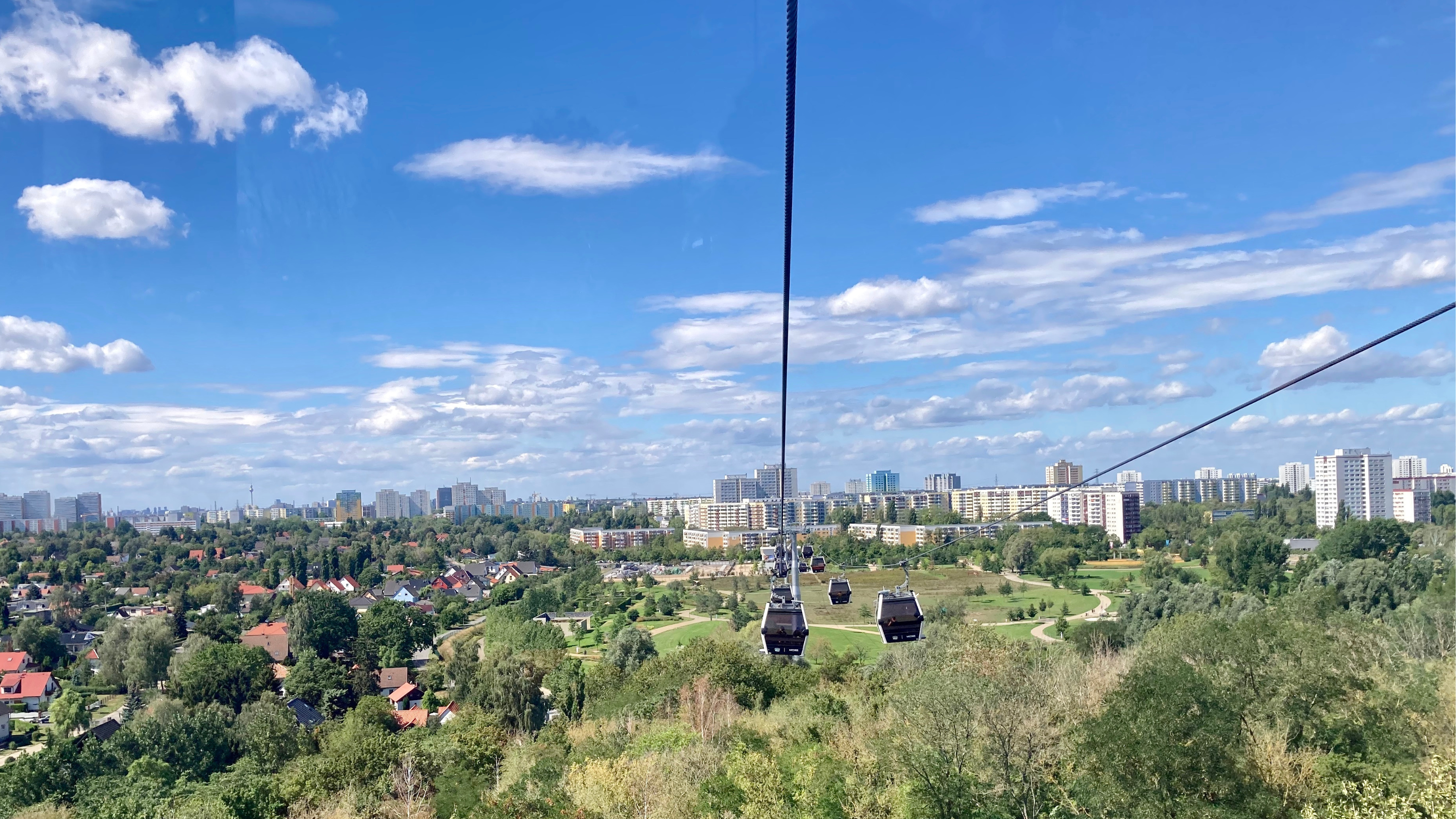 Panoramablick aus der Gondel einer Seilbahn über dem grünen Kienberg. Im Hintergrund sind Hochhäuser und der Fernsehturm zu sehen.